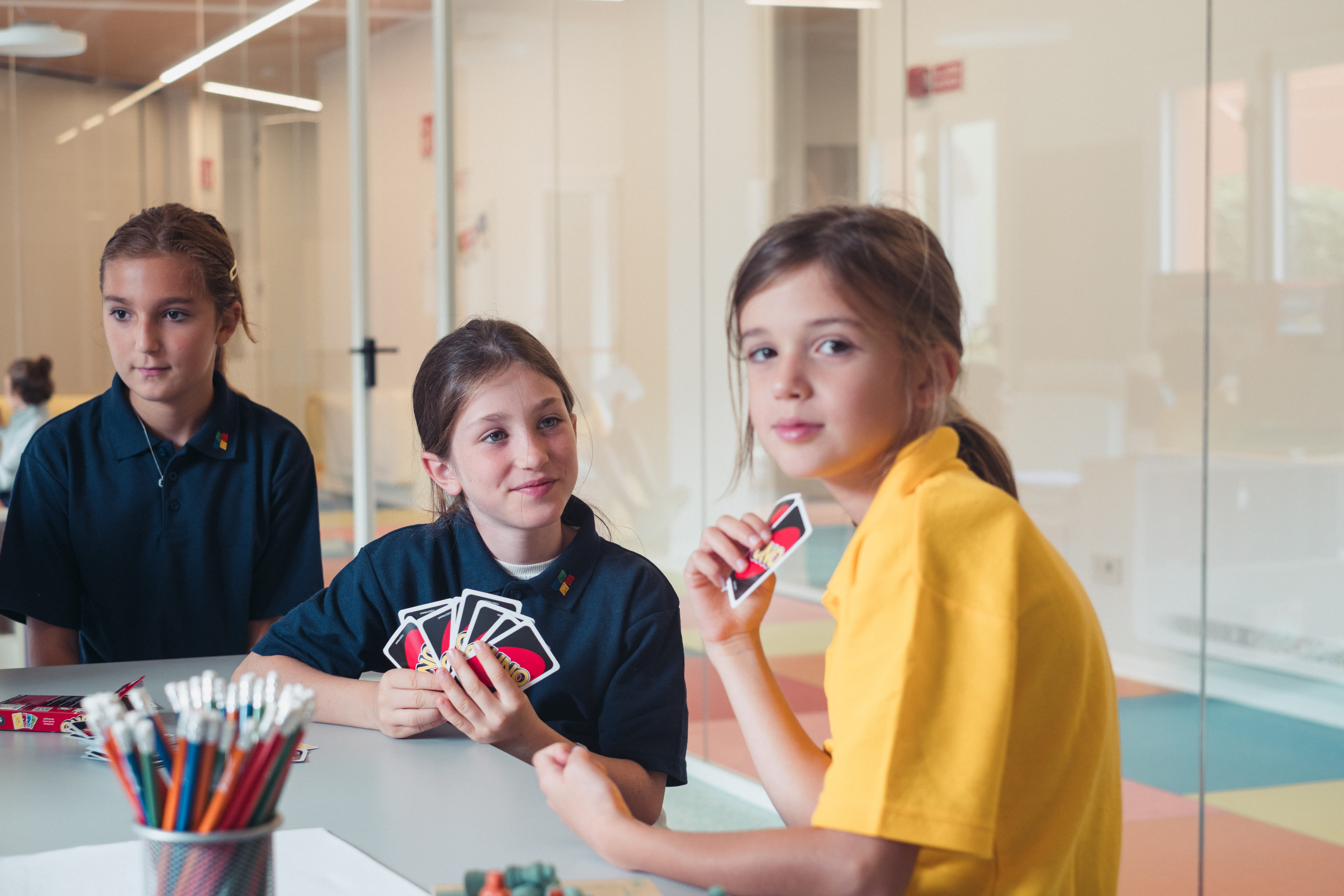 Three children are sitting together in the school campus, playing the card game Uno and smiling as they enjoy their break.