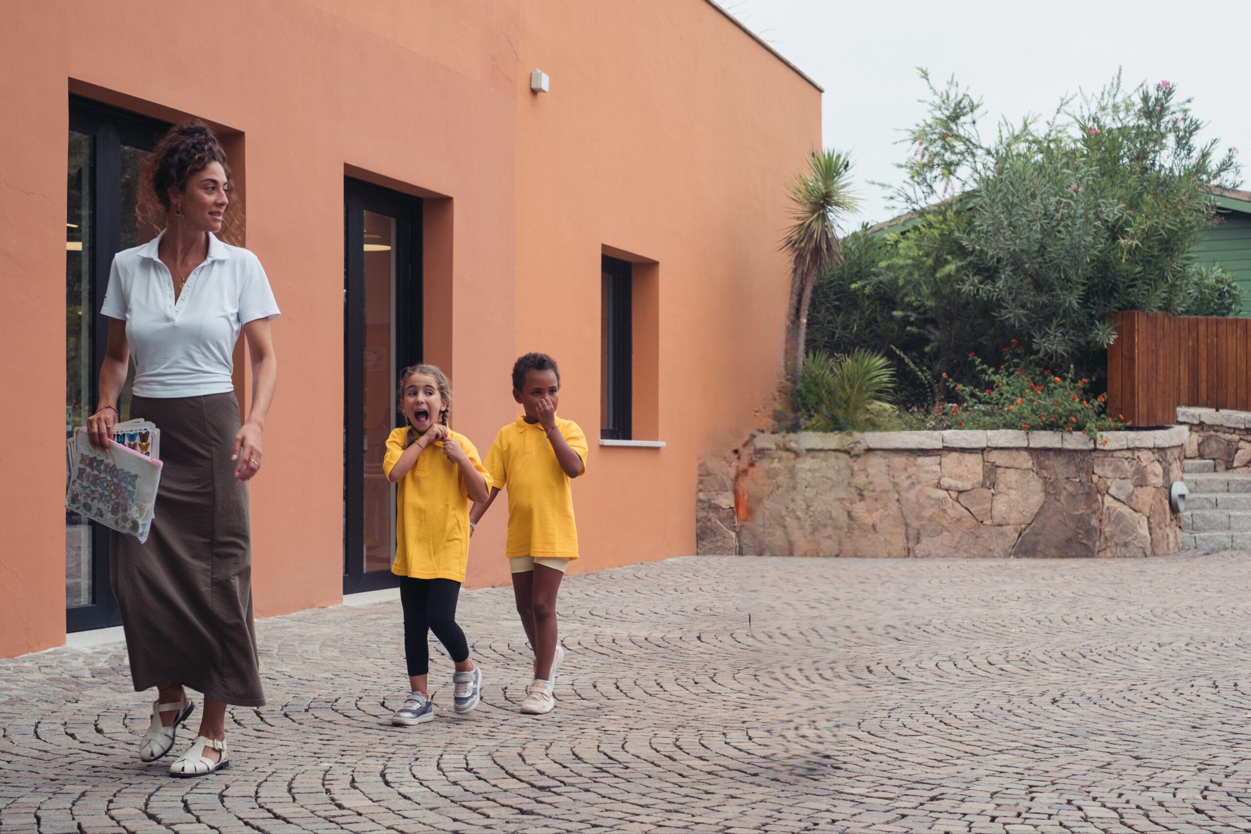 A teacher stands with a group of young children in a school courtyard. The children are wearing bright shirts and walking or running on a paved area near a modern orange school building, suggesting an outdoor lesson or activity. Shrubs and stone walls decorate the background, contributing to a safe and pleasant learning environment.
